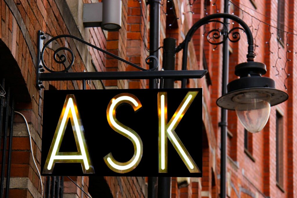 A sign hanging from a brick building with an old fashioned street light attached at the end. The sign has a black service with the word ASK in white and outlined in gold large letters